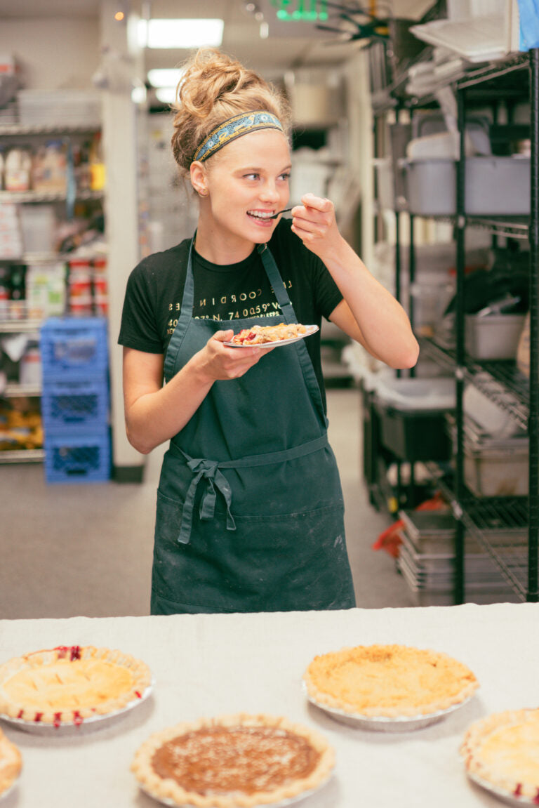 Woman in a kitchen eating a pie with pies on a counter in front of her.